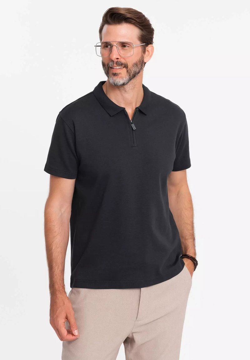 Černé polo triko s polovičním zipem, krátkým rukávem a měkkou texturou. Nosené přes světlé kalhoty, s volným střihem.