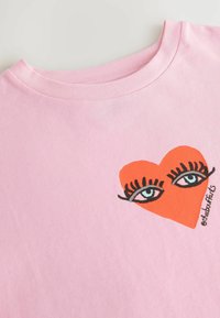 Rosa bomullströja med en grafik av stora blå ögon med svarta ögonfransar och en orange hjärtform. Crew neck-design, mjuk textur.