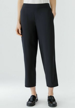 Zwarte op maat gemaakte enkellange broek gedragen met zwarte loafers, getoond op een persoon die staat tegen een eenvoudige lichte achtergrond.