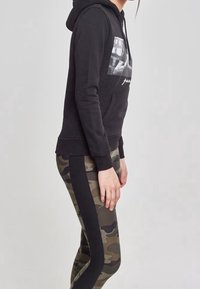 Felpa nera con grafica frontale e tasca a marsupio, abbinata a leggings camouflage oliva con pannelli laterali neri.