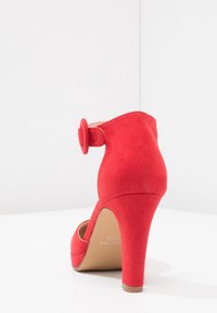 Červená semišová lodička na vysokém podpatku s zaoblenou špičkou a zakřiveným kotníkovým páskem s kruhovým akcentem. Hladký povrch a elegantní design.