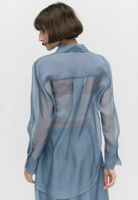 Vaaleansininen läpinäkyvä paita, jossa on kaulus, pitkät hihat ja rento malli. Huomionarvoista läpinäkyvää yksityiskohtaa selkäpuolella, joka korostaa kerroksellisuutta.