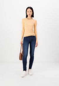 Lange mouw ribgebreide top in lichtoranje met kantdetails bij de halslijn, gecombineerd met donkerblauwe skinny jeans en witte sneakers. Bruine handtas.