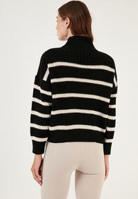 Zwarte en crèmekleurige gestreepte trui met een hoge hals, chunky brei-textuur en een cropped ontwerp. Heeft wijde mouwen en een ontspannen pasvorm.