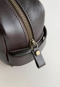 Geantă din piele maro închis cu formă rotunjită, finisaj texturat, fermoar auriu și o curea din piele cu detaliu metalic sub formă de nasture.