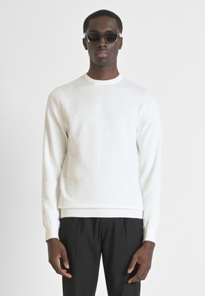 Maglione bianco dalla texture con collo girocollo, polsini e orlo a coste, abbinato a pantaloni neri. Modella con occhiali da sole neri.