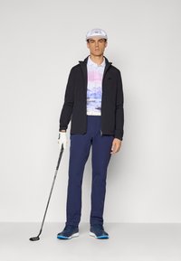 Férfi fehér sapkában, fekete dzsekiben, festői mintás pólóban, kék nadrágban és golfcipőben áll, kezében golfütőt tart, egy kesztyűs kézzel, sima háttér előtt.