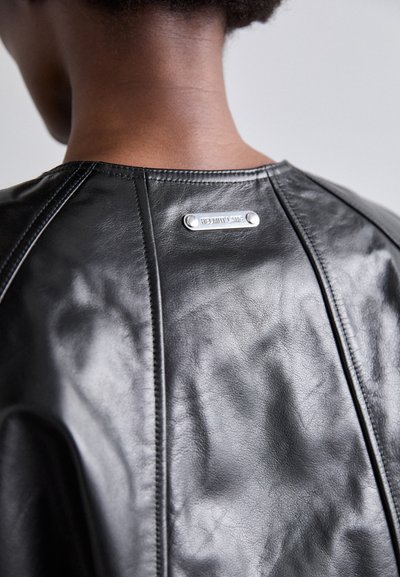 Fekete bőrkabát, lekerekített nyakkal, finom varrási részletekkel és a hátsó részen fémből készült címkével, amelyen domborított felirat található.