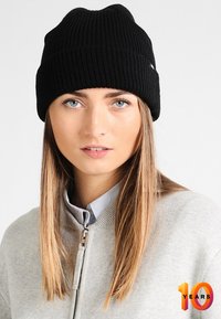 Beanie a coste nera con un design testurizzato, indossato da una persona in una giacca grigia chiara con zip sopra una camicia con colletto.