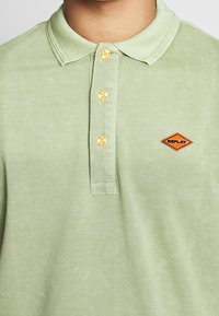 Jasnozielona koszulka polo z teksturowanej tkaniny, z trójprzyciskowym zapięciem i pomarańczowym diamentowym logo po lewej stronie na klatce piersiowej.
