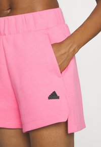 Vaaleanpunaiset urheilushortsit pehmeästä kankaasta, joissa on joustava vyötärönauha, sivutaskut ja musta Adidas-logo vasemmassa lahkeessa.