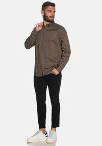 Camicia marrone da uomo con bottoni e maniche lunghe, abbinata a pantaloni neri slim-fit e sneakers bianche. Tessuto liscio, vestibilità casual.