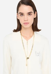 Cardigan zip-up bianco sporco con colletto e un logo decorato con strass. Una collana in catena dorata pende attorno al collo, aggiungendo un tocco di dettaglio.