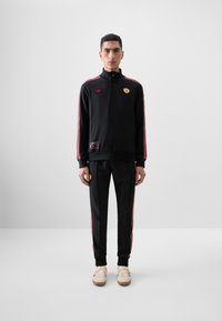 Black tracksuit with red side stripes, zippered jacket, and embroidered logos; paired with cream sneakers and fitted pants.