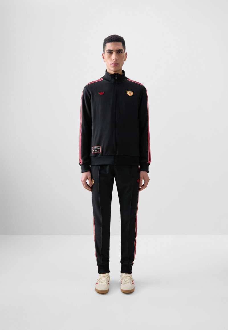 Black tracksuit with red side stripes, zippered jacket, and embroidered logos; paired with cream sneakers and fitted pants.