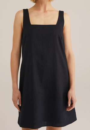 Vrouw draagt een mouwloze zwarte jurk met een vierkante halslijn, staand tegen een neutrale achtergrond.