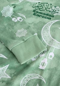 Grüner Pullover mit weißen Mustern von Mondsicheln, Laternen und floralen Designs. Der Text lautet "Ramadan Kareem." Rippenbündchen.