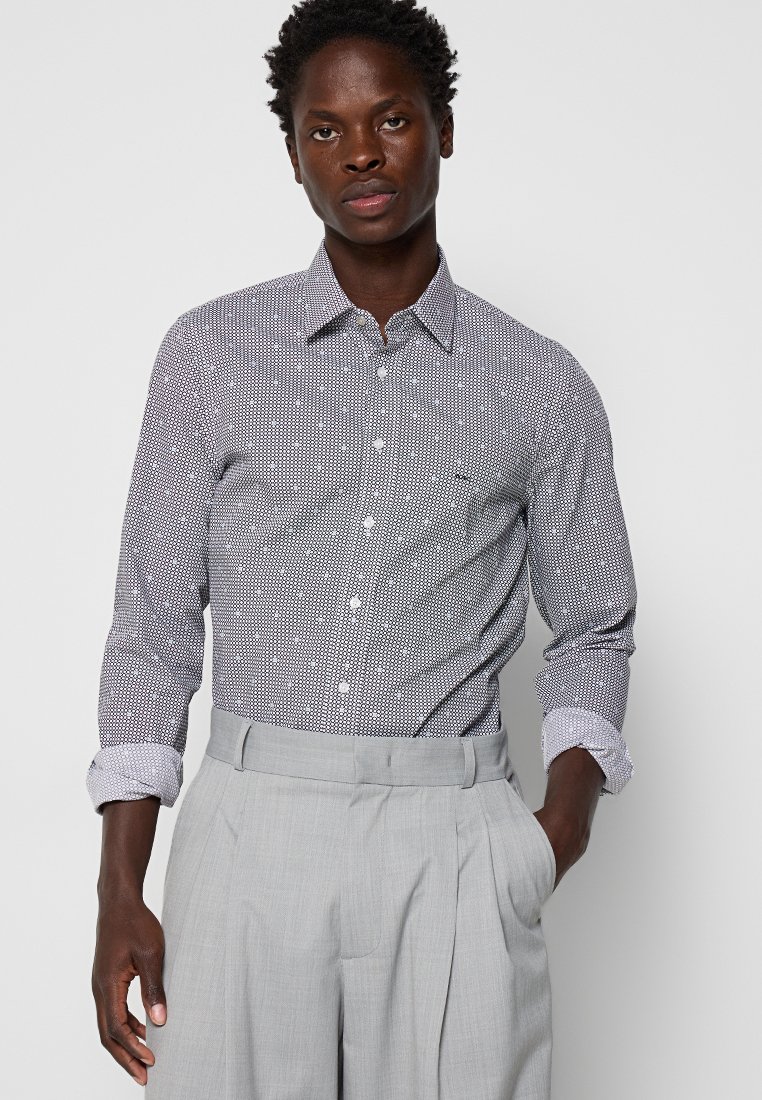 Camisa de botonadura con un patrón en blanco y negro, con cuello punta y mangas enrolladas. Llevada con pantalones de tejido gris claro con pliegues.