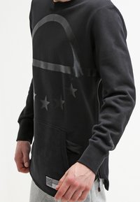 Svart sweatshirt med rund hals, med en grafisk design av stjärnor och en böjd linje över bröstet. Inkluderar en framficka och sidogångjärn.