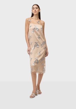 Femme portant une robe midi beige sans manches avec un voile transparent et des embellissements floraux argentés, assortie à des talons ouverts argentés.