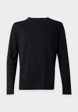Jeune homme portant un t-shirt noir à manches longues, un jean bleu clair à jambes larges et des baskets noires, debout devant un fond uni gris clair.