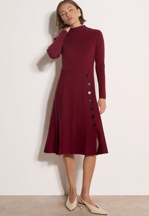 Mujer que lleva un vestido midi burdeos de manga larga con botones laterales y zapatos slingback beige de tacón bajo, de pie contra una pared lisa.