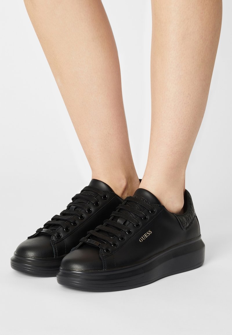 Baskets en cuir noir avec semelles plates, bouts ronds et fermeture à lacets. Présentent des détails embossés et un logo sur le côté.