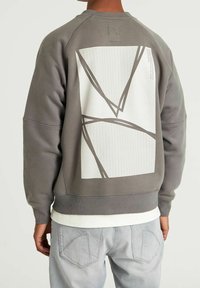 Sweat-shirt en coton gris avec un motif géométrique blanc dans le dos, doté d'un ourlet et de poignets côtelés. Portez par-dessus une chemise blanche.