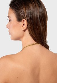 Gold necklace with a textured design, featuring a central clasp. Long, straight brown hair partially visible. Smooth skin tone in background.