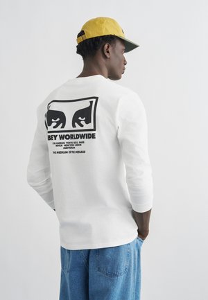 Mężczyzna w żółtej czapce, ubrany w białą koszulę z długim rękawem z czarnym nadrukiem oczu i napisem "OBEY WORLDWIDE" na plecach, do tego niebieskie dżinsy.