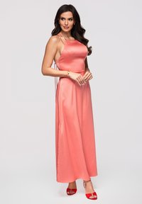Femme aux longs cheveux foncés portant une robe en satin corail rose à col halter et des talons rouges ouverts, debout devant un fond uni.