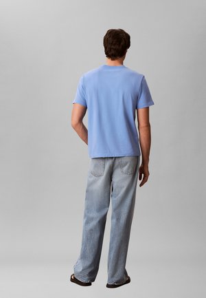 Mężczyzna widziany od tyłu, ubrany w jasnoniebieską koszulę z krótkim rękawem, luźne jasne dżinsy i czarne sandały, stojący na tle szarego tła.