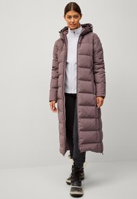 Cappotto lungo e imbottito color malva con cappuccio, dotato di chiusura con zip e polsini elastici. Indossato sopra una felpa grigia chiara con zip, abbinato a pantaloni neri e stivali.