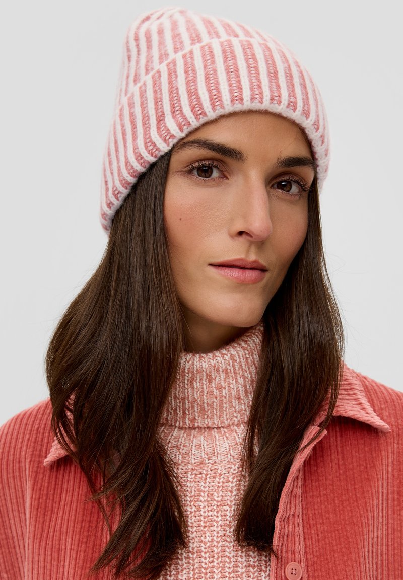 Bonnet en tricot rayé rose et blanc avec une coupe ajustée. Texture douce. Porté avec un pull rose à col montant et une veste en velours côtelé assortie.