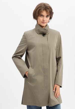 Junge Person mit kurzen braunen Haaren, die einen hellbraunen, geknöpften Mantel trägt und mit einer Hand in der Tasche vor einem schlichten Hintergrund steht.