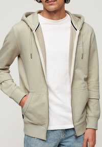 Felpa zip con cappuccio foderato di un grigio chiaro, con tasche frontali e polsini a coste. Indossata sopra una t-shirt bianca, presenta un design semplice e un branding minimale.