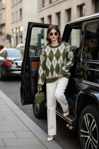 Femme portant un pull à losanges vert et blanc et un pantalon blanc sort d'un SUV noir dans une rue de la ville, tenant un petit sac à main vert.