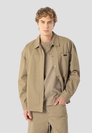 Vaaleanruskea napillinen paita, joka on valmistettu pehmeästä kankaasta, jossa on rintatasku ja rento istuvuus. Paita on käytetty beigeä t-paitaa vasten.