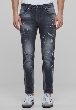 Mann trägt dunkelgraue, abgenutzte Jeans mit Farbspritzern und weiße Turnschuhe mit schwarzen Streifen, steht vor einem schlichten Hintergrund.