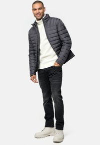 Chaqueta puffer gris con cuello alto, suéter de punto blanco, jeans negros y botas de tobillo beige. Presenta un diseño acolchado horizontal y cierre de cremallera.