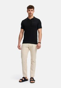 Black knitted short-sleeve polo shirt with pocket, paired with beige trousers and dark sandals with straps. Simple, modern design.