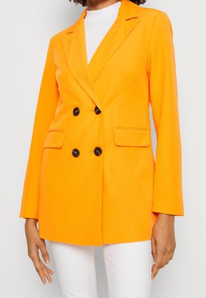 Vrouw draagt een feloranje dubbelgeknoopt blazer met zwarte knopen over een wit topje en witte broek, staand tegen een effen achtergrond.