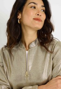 Veste bomber métallique dorée avec fermeture éclair à l'avant, col arrondi et poignets côtelés. Texture lisse avec un léger éclat. Associée à des boucles d'oreilles créoles.