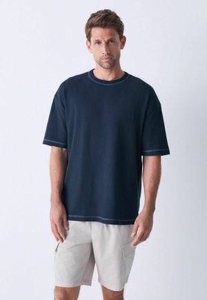 T-shirt bleu marine à manches courtes en matériau texturé, avec un col rond et une couture contrastante. Porté avec un short beige clair.