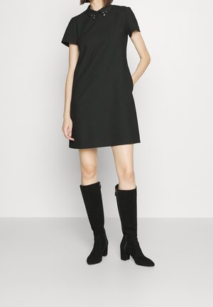 Nainen, joka pukeutunut lyhythihaiseen mustaan mekkoon, jossa on niitattu kaulus, ja mustiin polven korkeisiin korkosaappaisiin, seisoo valkoista taustaa vasten.