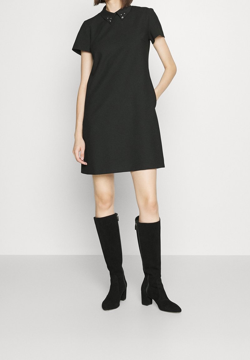 Vrouw draagt een korte zwarte jurk met een met studs versierde kraag en kniehoge zwarte laarzen met hak, staand tegen een witte achtergrond.