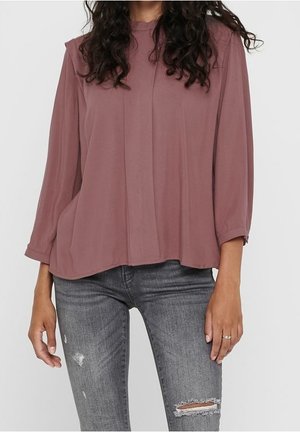 Femme portant un chemisier ample mauve à manches longues et un jean gris déchiré avec des déchirures sur le genou droit et la cuisse gauche.