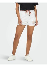 Beli traper šortsi z raztrganim robom, sprednjimi raztrganinami in barvnim vzorcem vezalk na pasu, v kombinaciji z belimi supergami.