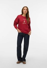 Rdeč pulover z besedilom "SEASON'S GREETINGS" in grafiko drevesa. Kombiniran s temno modrimi zvončastimi kavbojkami in rjavimi čevlji. Priložnostni videz.