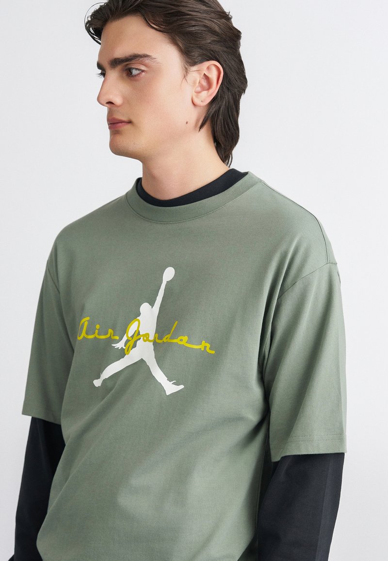 Ελιάς πράσινη μπλούζα T-shirt με στρογγυλή λαιμόκοψη, με λογότυπο Jordan σε λευκό χρώμα και κίτρινο κείμενο "Air Jordan". Το ύφασμα φαίνεται απαλό, με χαλαρή εφαρμογή.